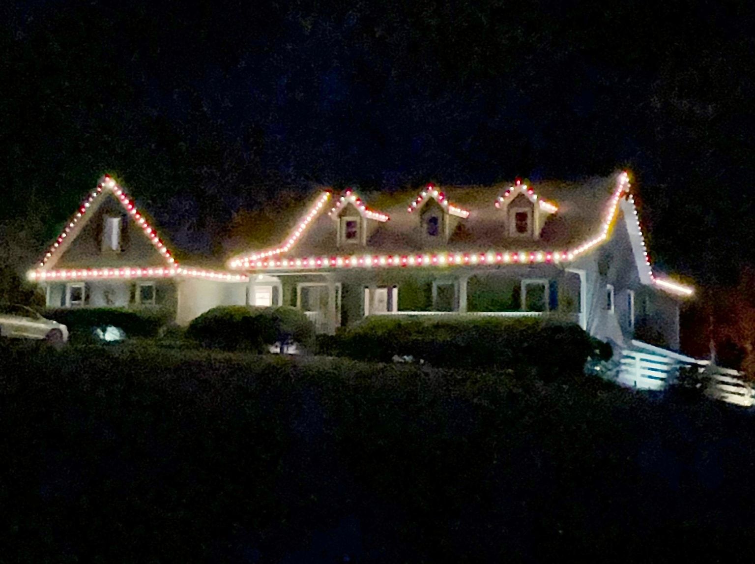 Red and white roofline Christmas lights outlining the gables and three dormers of a hillside home
